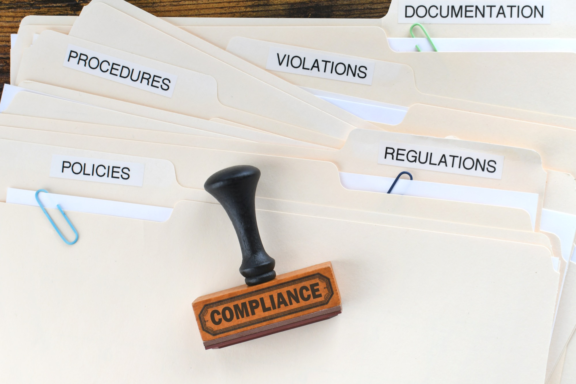 A compliance stamp resting on folders labeled documentation, regulations, and procedures
